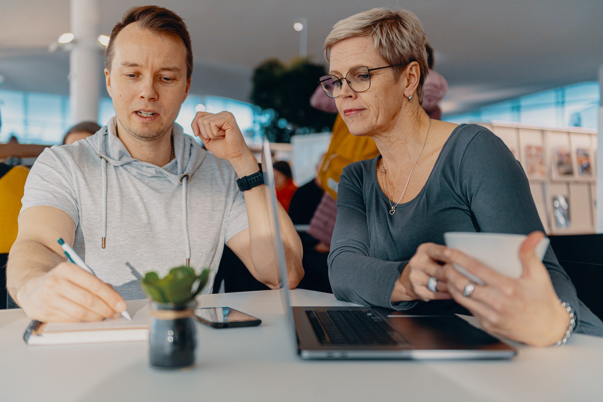 Mies kirjoittaa muistivihkoon. Nainen katsoo vieressä. Pöydällä on kannettava tietokone.