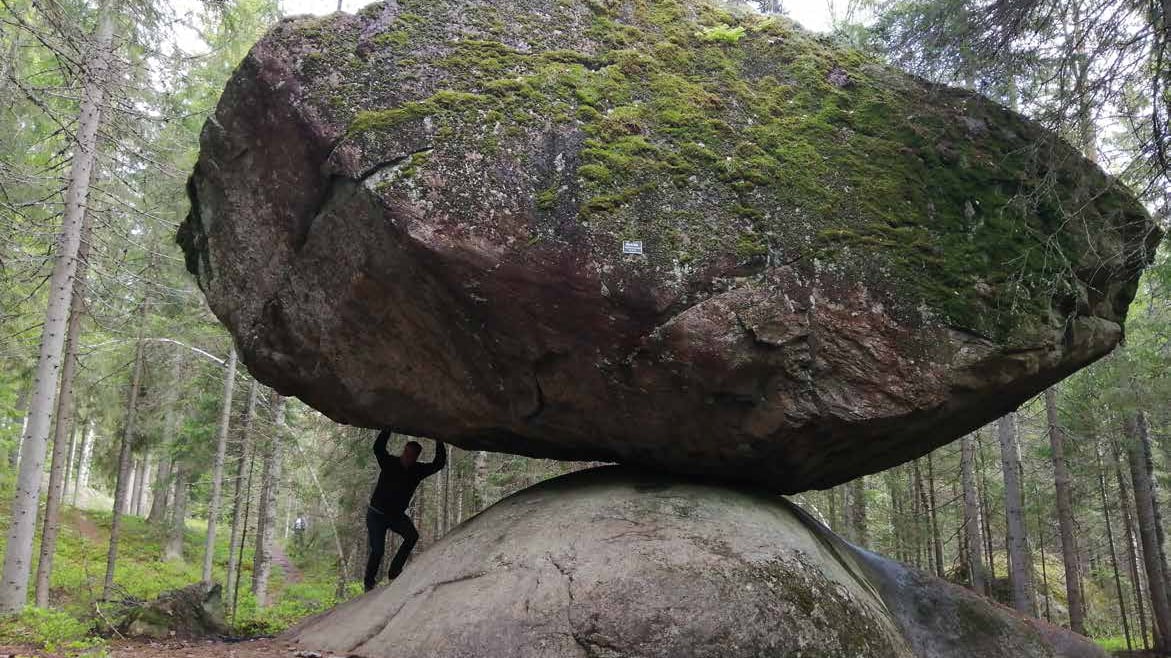 Suuri siirtolohkare, joka on maata vasten vain pieneltä kulmaltaan. Kiven alla on mies, joka näyttää ikään kuin pitelevän lohkaretta ilmassa.